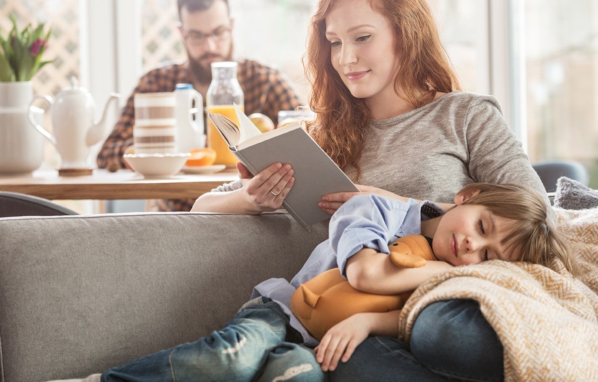 child cuddling on mother's lap while she reads a book on the couch and dad is in the background at the kitchen table